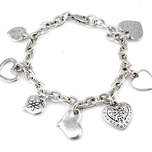 May include: A silver charm bracelet with a chain link design and multiple heart-shaped charms. The charms include a heart with a floral design, a heart with a star, a plain heart, and a heart with a hammered texture.