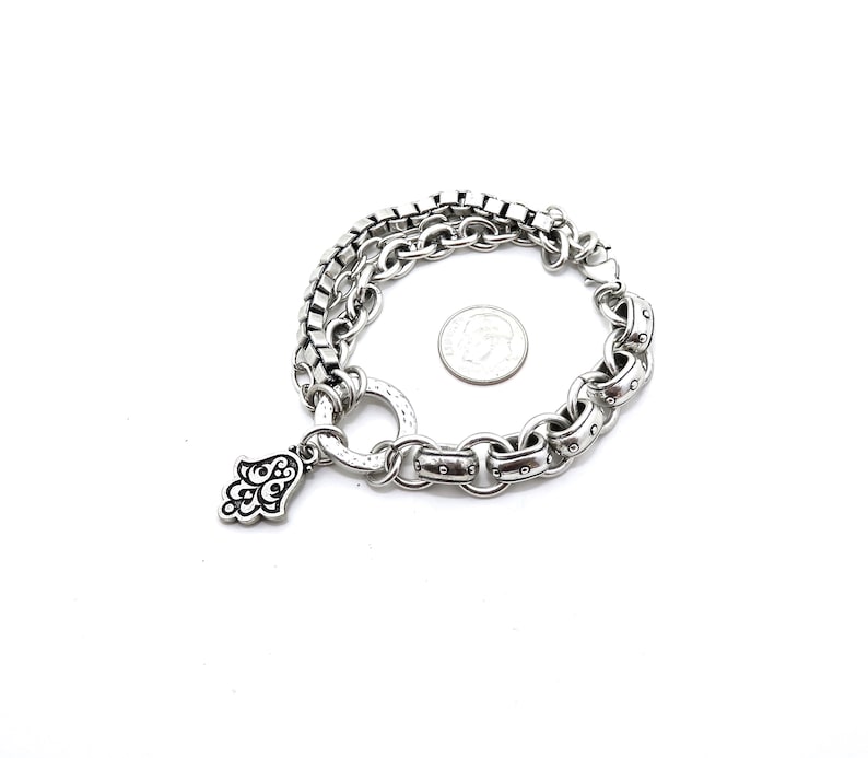 May include: Silver-toned bracelet featuring a layered design with a square chain and a larger link chain. A small Hamsa hand charm hangs from the bracelet. A US quarter is included for size reference. The bracelet is on a white background.