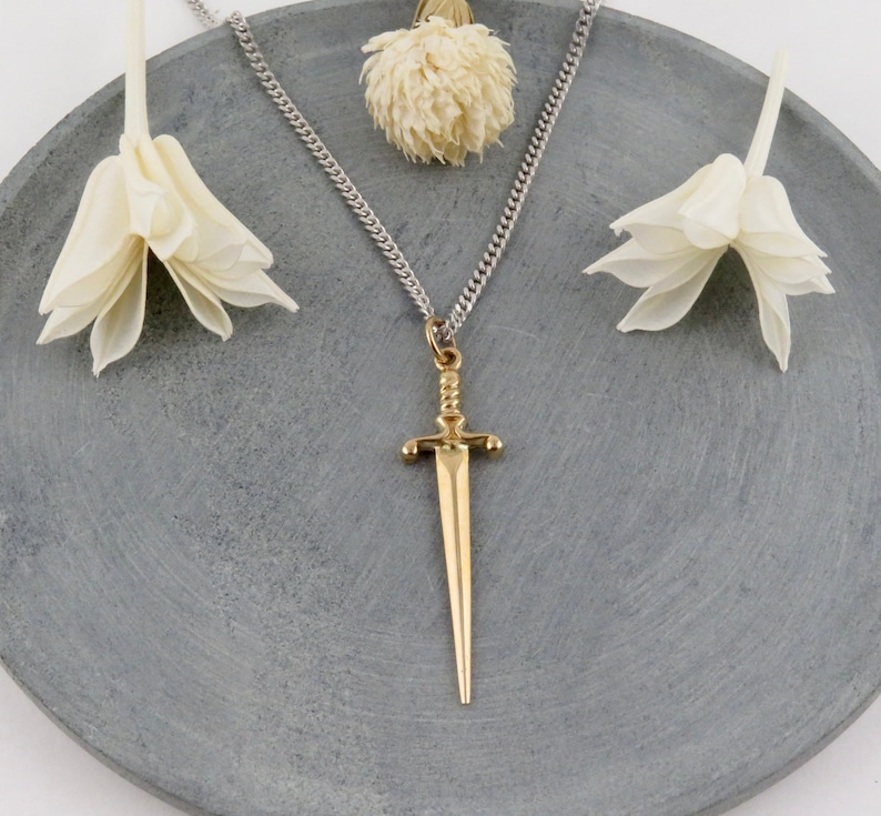 Peut inclure: Un collier en cha&icirc;ne argent&eacute;e avec un pendentif en forme d'&eacute;p&eacute;e dor&eacute;e. Le pendentif est un design simple et stylis&eacute; avec une pointe pointue. Le collier est pr&eacute;sent&eacute; sur une surface grise avec des fleurs blanches.