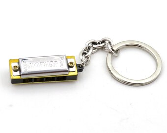 May include: A small gold harmonica keychain with a silver metal body and a silver chain with a silver key ring. The harmonica has the word "HOHNER" printed on it.