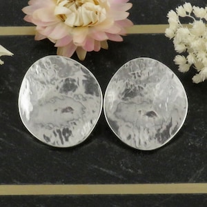 May include: A pair of silver-toned, hammered-metal disc earrings. The earrings have an irregular, oval shape and a textured surface. The background features a black surface with a gold stripe and dried flowers.