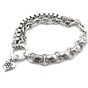 May include: Silver-toned bracelet featuring a mix of chain styles, including box chain and round links. A heart-shaped charm with a starburst design hangs from the bracelet. The bracelet is set against a white background.