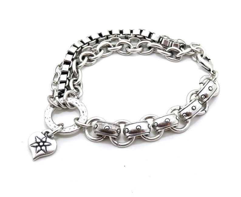 May include: Silver-toned bracelet featuring a mix of chain styles, including box chain and round links. A heart-shaped charm with a starburst design hangs from the bracelet. The bracelet is set against a white background.