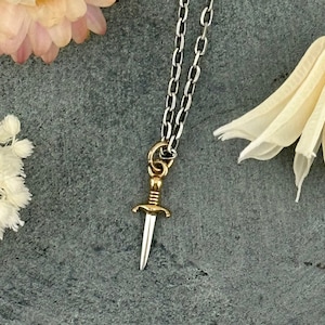 May include: A silver chain necklace with a gold dagger pendant. The dagger is detailed with a twisted handle and a sharp point.
