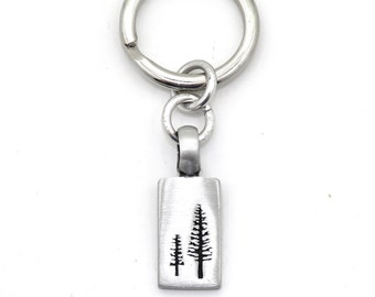 May include: A silver keychain with a rectangular charm featuring a silhouette of two pine trees. The charm is attached to a silver key ring.