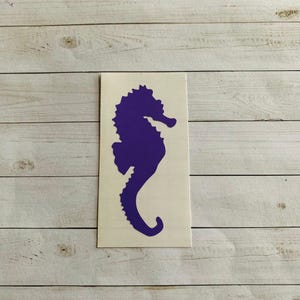 May include: A purple seahorse decal on a white rectangular background. The seahorse silhouette is a solid, vibrant purple, with a detailed, textured appearance. The decal is placed against a light wood-grain background.