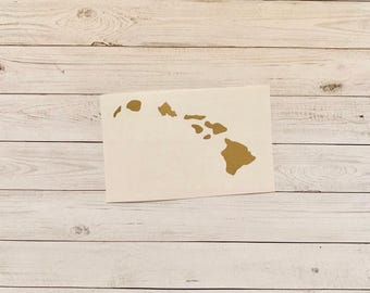 Könnte beinhalten: Ein rechteckiger, cremefarbener Aufkleber mit einer goldfarbenen Umrisszeichnung der hawaiianischen Inseln. Der Aufkleber liegt auf einer hellen Holzoberfläche mit horizontalen Brettern.