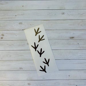 May include: A white rectangular sticker featuring five black bird footprints. The footprints are evenly spaced and angled, creating a visual pattern. The sticker is placed on a light-colored, wooden surface.