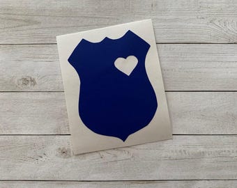 May include: A blue shield-shaped sticker with a white heart cutout. The sticker is on a white background and placed on a light wood grain surface. The shield has a curved top and a pointed bottom.