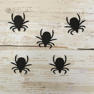 May include: Five black paper spider cutouts on a white wood background.
