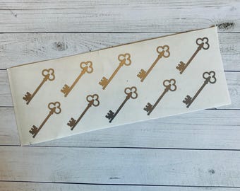 May include: A sheet of stickers with gold and bronze key designs. The keys are in rows, with metallic gold and bronze shades. The stickers are on a white rectangular sheet, against a light wood-grain background.