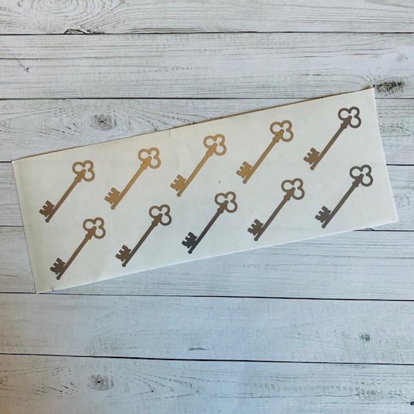 May include: A sheet of stickers with gold and bronze key designs. The keys are in rows, with metallic gold and bronze shades. The stickers are on a white rectangular sheet, against a light wood-grain background.