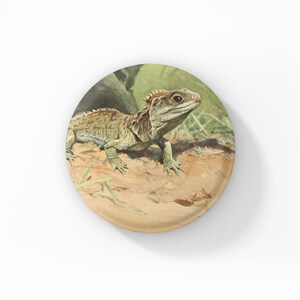 May include: A round decorative item featuring a detailed illustration of a lizard. The lizard is depicted in shades of green and brown, resting on a sandy surface with green foliage in the background. The item has a white background.