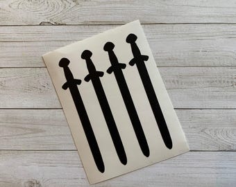 May include: A white sticker featuring five black sword silhouettes. The swords are evenly spaced and have a classic design, with a crossguard and a rounded pommel. The sticker is on a light wood grain surface.