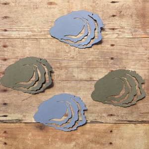 May include: Four paper cutouts of oyster shells in two different colors. Two are light blue and two are dark green. The cutouts are arranged in a square pattern on a wooden surface.