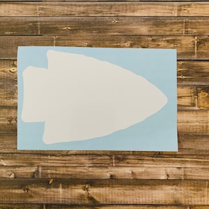 May include: A white arrowhead shape on a light blue background. The arrowhead is centered on the blue rectangle, which is placed on a wooden surface. The image is a top-down view.