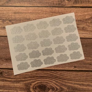 May include: A sheet of silver glitter cloud stickers. Each cloud is a puffy shape with a sparkly texture. The stickers are arranged in rows and columns on a white background. The background is a wooden surface.
