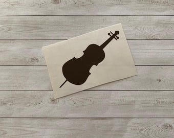 May include: A brown cello silhouette on a white rectangular sticker. The sticker is placed on a light-colored wooden surface. The cello design features the instrument's body, neck, and a single endpin.