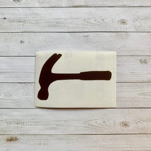 May include: A dark brown hammer sticker on a white background. The hammer is a silhouette, with a curved claw and a long handle. The sticker is rectangular and sits on a light wood surface.