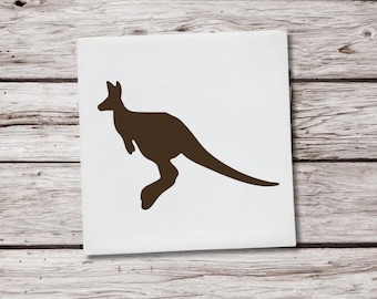 May include: A brown silhouette of a kangaroo on a white square. The kangaroo is depicted in a side profile, with its tail extended and its legs in a jumping position. The background is a light-colored wooden surface.
