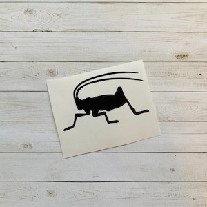 May include: A black silhouette of an insect with long antennae and legs, positioned on a white rectangular sticker. The sticker is set against a backdrop of light-colored, horizontal wooden planks.