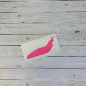 May include: A bright pink slug sticker on a white background. The sticker is a solid color and has a simple, cartoon-like design. The background is a light-colored, wooden surface.