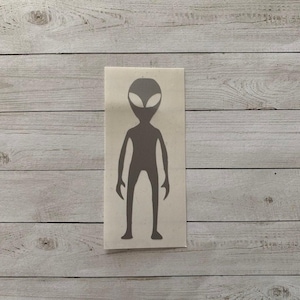 May include: A gray alien sticker on a rectangular white background. The alien has a large head with oval eyes, a slender body, and long arms and legs. The sticker is placed on a light wood grain surface.