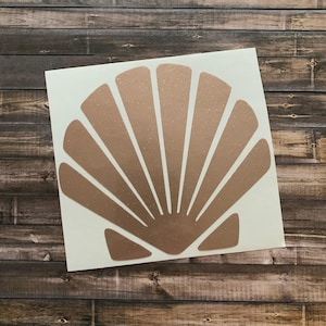 May include: A seashell-shaped sticker in a metallic rose gold color. The sticker is on a white background, with a wooden surface in the background. The seashell design features radiating lines, creating a fan-like pattern.
