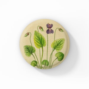 May include: Round, cream-colored decorative plate featuring a botanical illustration of a violet plant. The artwork showcases green leaves, purple flowers, and buds, set against a vintage-style background. A charming piece for home decor.