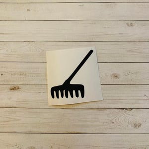 May include: A black rake graphic on a white square sticker. The sticker is placed on a light wood-grain surface. The rake has a long handle and multiple tines, suitable for gardening and yard work.