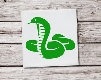 May include: A green snake illustration on a white square. The snake is stylized with a curved body and raised head, with a forked tongue. The background is a light wood grain.