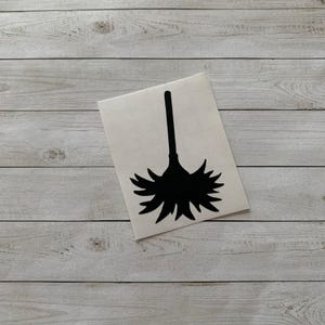 May include: A black silhouette of a broom on a white sticker. The broom has a long, straight handle and a flared, detailed brush head. The sticker is angled on a light gray wooden surface.