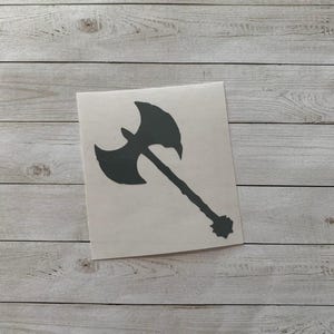 May include: A dark gray axe sticker on a white square. The axe has a double-bladed head and a long handle with a decorative end. The sticker is placed on a light gray wooden surface.