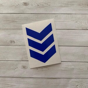 May include: A white sticker featuring three blue chevrons pointing downwards. The chevrons are evenly spaced and have a clean, modern design. The sticker is placed on a light-colored wooden surface.
