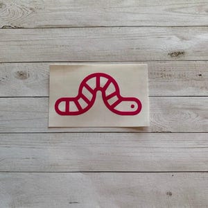 May include: A red worm sticker on a white background. The worm is stylized with segmented body and a small dot for an eye. The sticker is placed on a light wood grain surface.