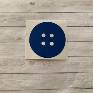 May include: A navy blue button sticker with four white holes. The sticker is round and has a simple, clean design. The sticker is on a white background with a wood grain pattern.