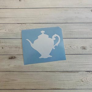May include: A white teapot silhouette on a light blue square, set against a backdrop of weathered, light-colored wooden planks. The teapot design features a curved spout, a looped handle, and a decorative lid. The overall aesthetic is simple and elegant.