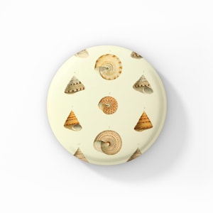 May include: A round, off-white button featuring a pattern of various seashells in shades of brown, tan, and cream. The seashells are illustrated in a vintage style, with detailed lines and shading. The button is a decorative item.