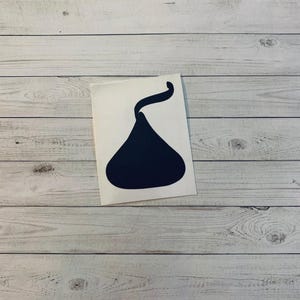 May include: A navy blue sticker in the shape of a Hershey's Kiss. The sticker is on a white background, set against a light wood grain surface.