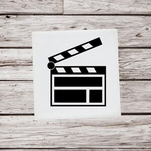May include: A black and white film clapperboard, a filmmaking tool, is displayed against a light wood background. The clapperboard is open, with a hinged top section and a rectangular body. The design is simple and graphic.