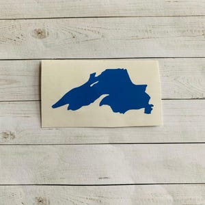 May include: A blue decal of Lake Superior, a large freshwater lake in North America. The decal is a solid blue color and is rectangular in shape. The decal is on a white background with a wood grain pattern.