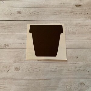 May include: A dark brown, stylized silhouette of a flower pot on a white background. The pot has a rectangular body with a slightly flared top. The sticker is placed on a light wood grain surface.
