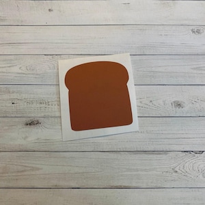 May include: A brown, bread-shaped sticker with a white border. The sticker is a solid color and is placed on a light-colored, wooden surface.