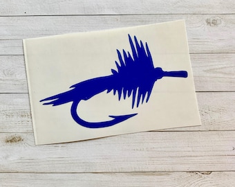 May include: A blue fly fishing lure sticker on a white background. The sticker features a detailed illustration of a fly fishing lure, with a curved hook and feathery details. The sticker is placed on a light wood grain surface.