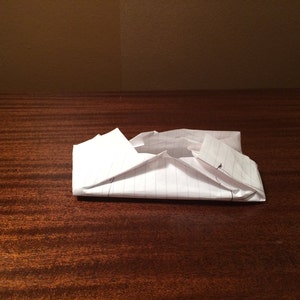 May include: A white piece of paper folded into a small, rectangular shape. The paper has lines on it and is sitting on a wooden surface.