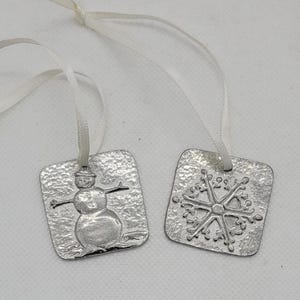 May include: Two silver metal ornaments with white ribbon. One ornament features a snowman with a hat and the other features a snowflake.