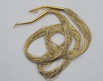 May include: Gold-toned necklace featuring multiple strands of fine wire. The necklace has a hook clasp and curved end pieces. The strands are gathered at each end, creating a flowing, layered effect. The jewelry is displayed on a white surface.