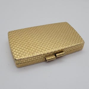 May include: A rectangular, gold-colored box with a textured, woven pattern. The box has a hinged lid and a clasp closure. The overall design is simple and elegant, suitable for small items.