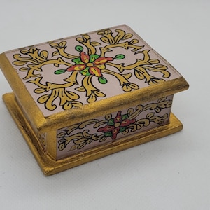 May include: A small, square decorative box with a hinged lid. The box is painted with a gold border and a pink background. The lid and sides feature a floral design in gold, green, red, and yellow.