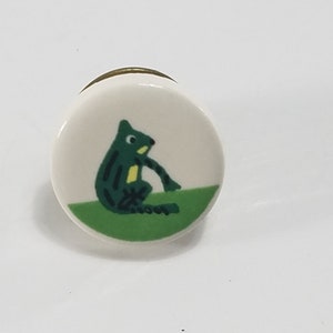 May include: A round ceramic knob with a green frog illustration on a green background. The knob has a gold rim.